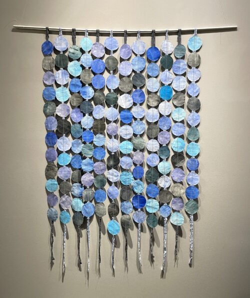A wall-hanging artwork with blue and black disks that are sewn to strips of fabric. Made from the novel, Black and Blue.