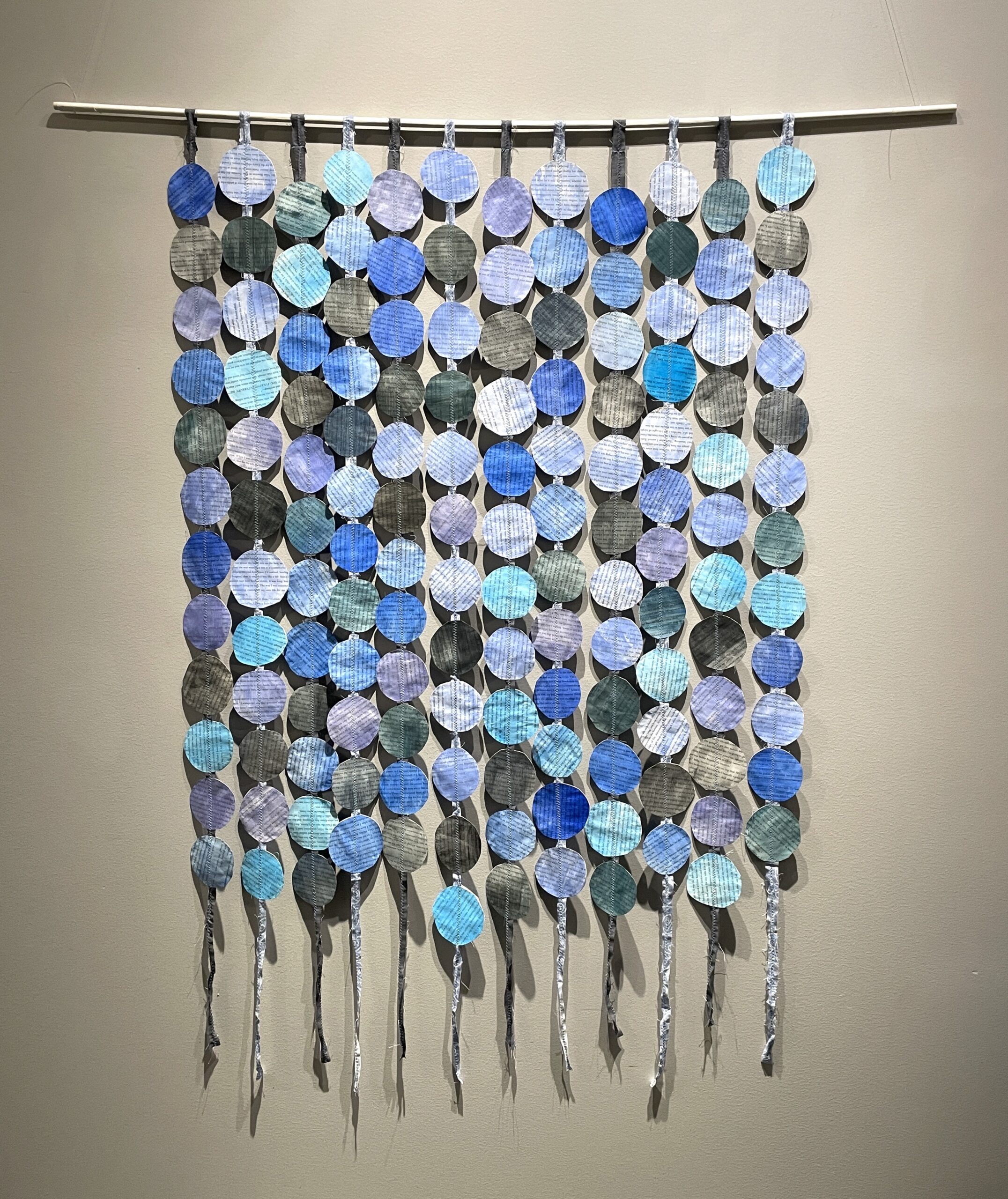 A wall-hanging artwork with blue and black disks that are sewn to strips of fabric. Made from the novel, Black and Blue.