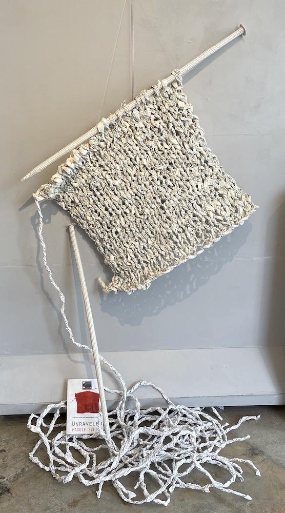 An artwork of book paper that has been twisted into yarn and then knitted with two giant knitting needles.