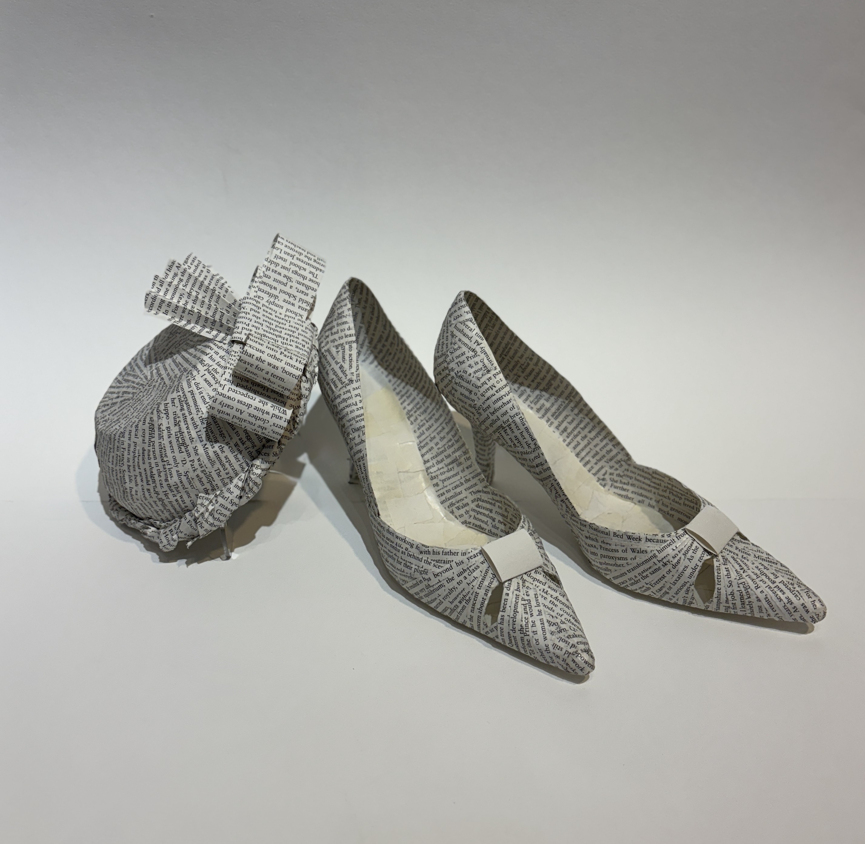 An artwork by Maggie Kerrigan that depicts a pair of low heeled shoes and a fashionable hat, the type that would have been worn by Princess Diana. The paper-cast items are made from the biography of the princess.