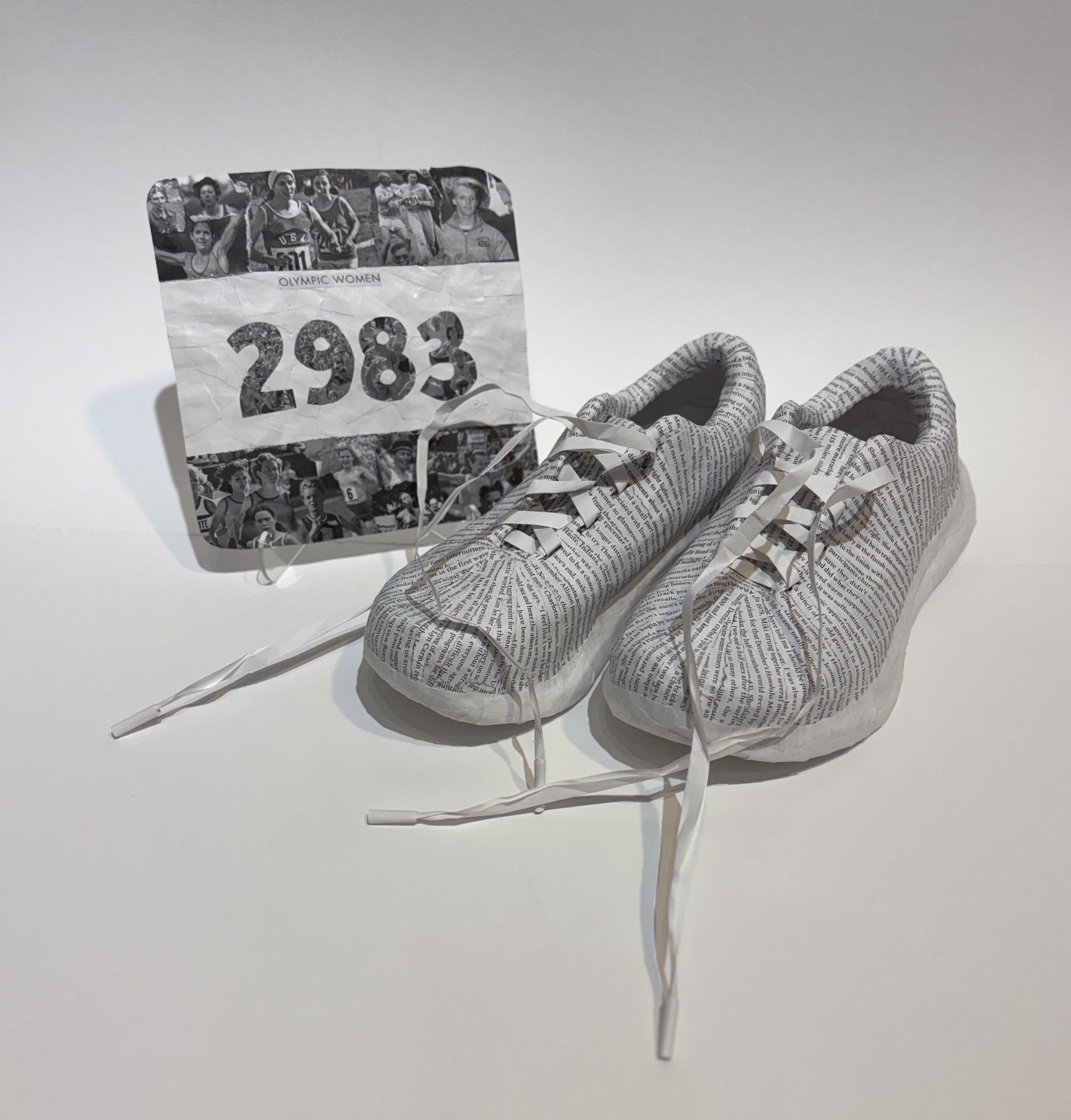 A pair of paper-cast running shoes and a runners bib created by artist Maggie Kerrigan. The shoes and bib are formed by paper from the book, First Ladies of Running.