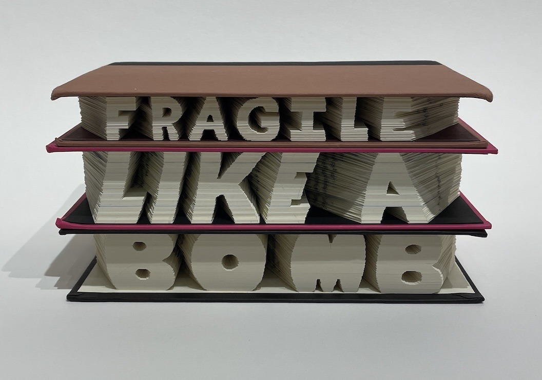 An artwork by Maggie Kerrigan with the words "Fragile Like A Bomb" carved into the edges of 3 books about Ruth Bader Ginsburg and Frida Kahlo.