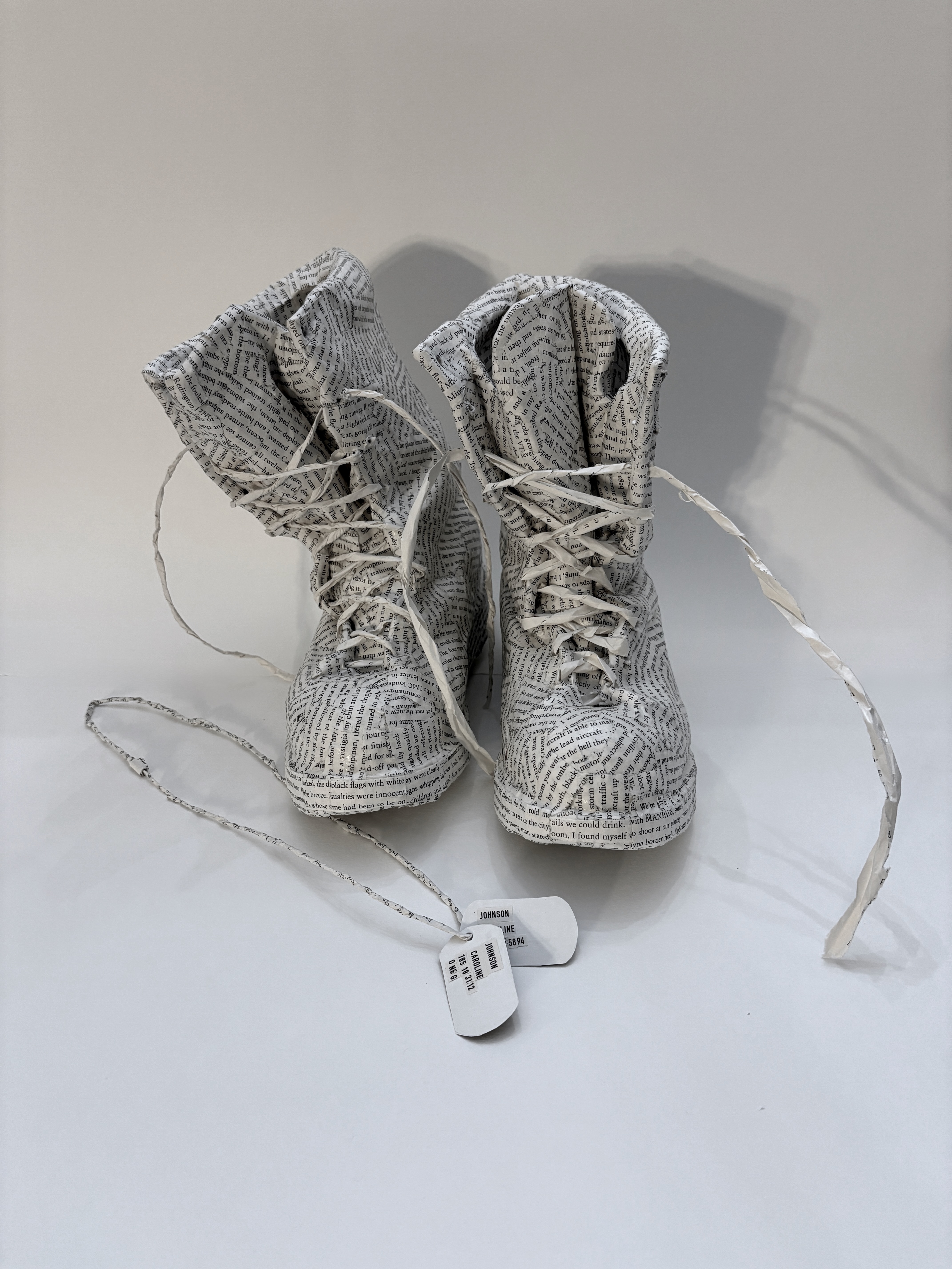 An artwork by Maggie Kerrigan. A pair of combat boots and set of dog tags that are made of paper from the book Jet Girl about a female fighter pilot.