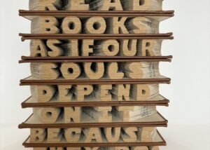 This altered book artwork is stack of 9 books hand-carved by Virginia Beach artist, Maggie Kerrigan, and reads as the sentence "We must read as if our souls depend on it, because they do!"